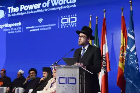 Rabbi Hofmeister stands at podium at KAICIID Power of Words Conference. Behind him sit 5 religious leaders.